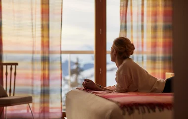 Mulher deitada na cama do quarto, observando a paisagem pela janela.
