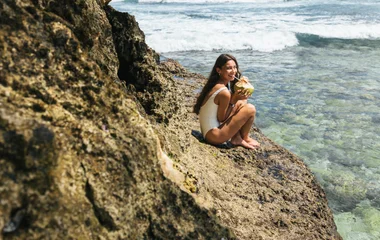 Una mujer bebe agua de coco sentada en una roca en la playa.