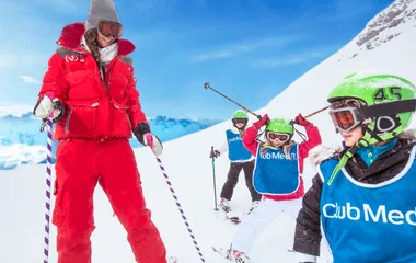 Instrutor de esqui vestido de vermelho e ao lado direito três crianças com coletes e capacetes de esqui verdes. Ao fundo a neve.