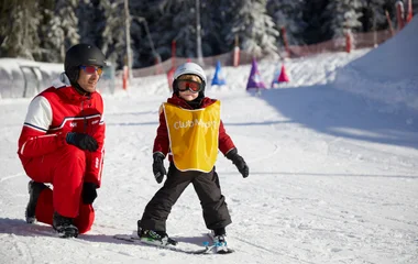 Criança pequena usando equipamentos de esqui e uma traje amarelo com o logo do Club Med é supervisionada por um adulto enquanto esquia na neve.