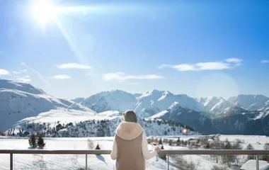 Mulher se debruça em varanda aberta com drink na mão enquanto admira paisagem de neve