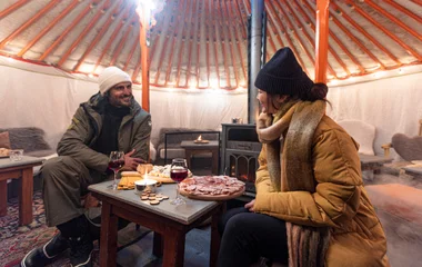 Casal se olhando sentados em uma mesa com petiscos e vinho.