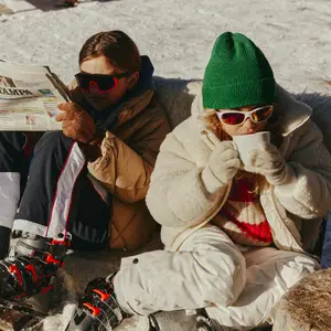 Criança toma chocolate quente sentada na neve e usa gorro verde e óculos escuros
