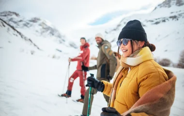 Mujer con gorro, anteojos de sol, campera de nieve color mostaza y equipo de esquí, caminando por la nieve.