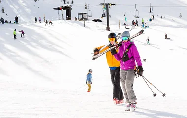 Casal andando e carregando com os equipamentos de esqui.