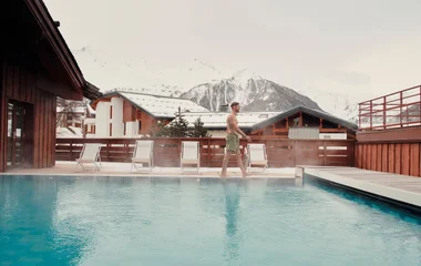 Homem andando na borda de piscina aquecida com vista para as montanhas nevadas ao fundo da foto