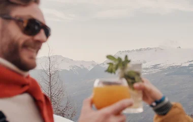 um casal brindando com drinks, e ao fundo montanhas cobertas de neve