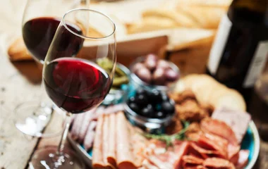 Duas taças de vinho com tábua de frios ao fundo.