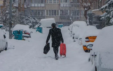 Vista de um homem de costas, vestindo um casaco preta e carregando uma mala vermelha, enquanto neva