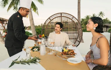 Casal trajando branco e com drinks na mão adornados por uma paisagem de por do sol na praia.