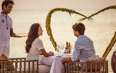 Casal sentado de frente para o mar sendo servidos por um garçom durante o por do sol e um arranjo floral de coração ao fundo.