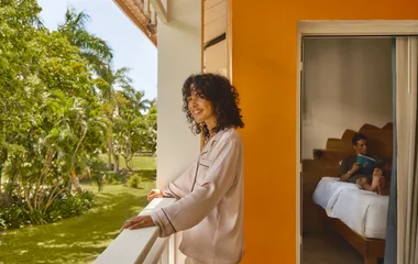 Mulher na varanda enquanto admira a vista e homem deitado na cama lendo um livro