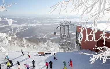 Vista panorâmica de montanha nevada com teleférico ao fundo, pessoas com equipamentos de esqui e árvores congeladas