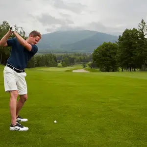 Um homem praticando golfe se preparando para acertar a bola com o taco levantado em um cenário verde sinuoso com árvores e montanhas