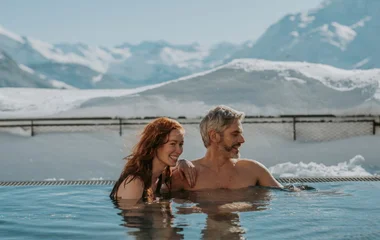 Casal em piscina aquecida com paisagem de neve ao fundo.